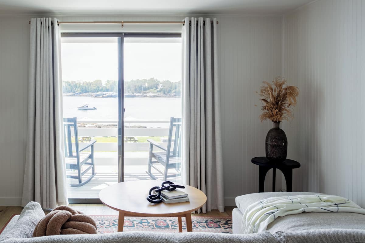 Ein Zimmer mit Blick auf ein Gewässer, ein Tisch mit Büchern und einer Sonnenbrille, ein Stuhl, eine Couch und eine Vase mit getrockneten Blumen.