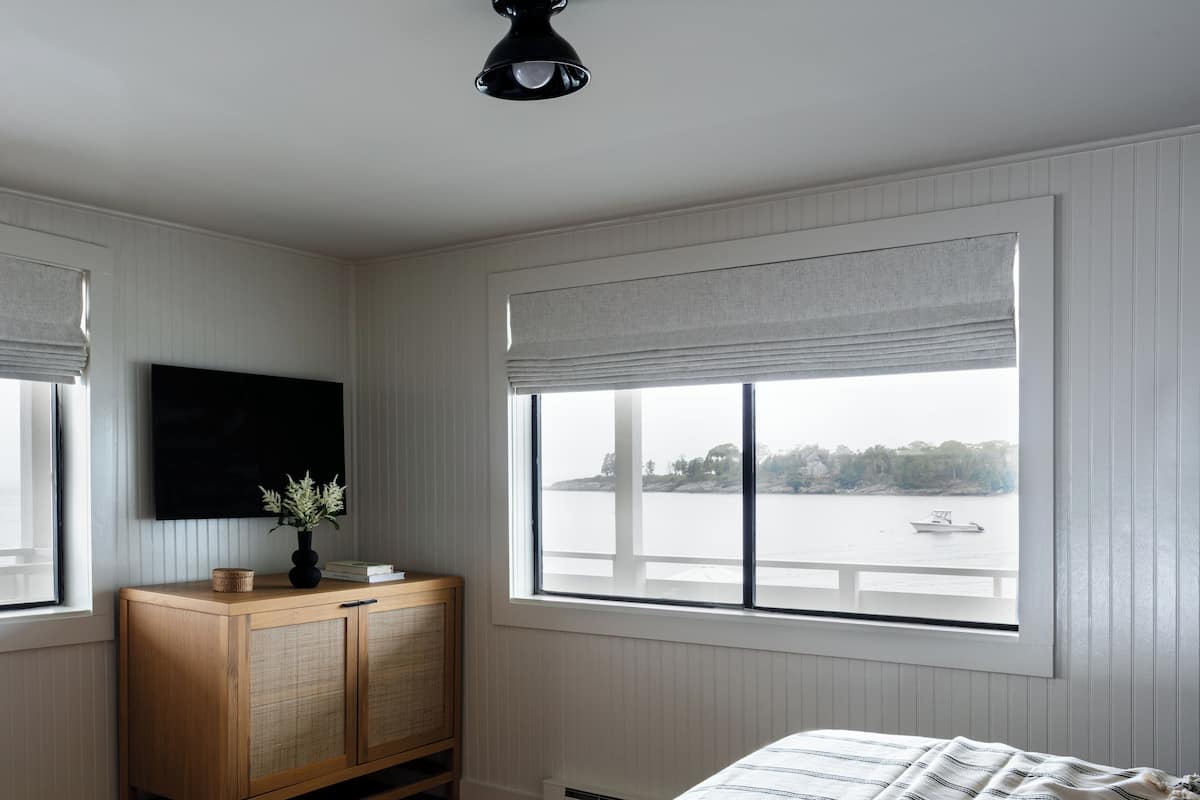 Ein Schlafzimmer mit einem Bett, einem Holzschrank, einem Fernseher und großen Fenstern mit Blick auf ein Gewässer.
