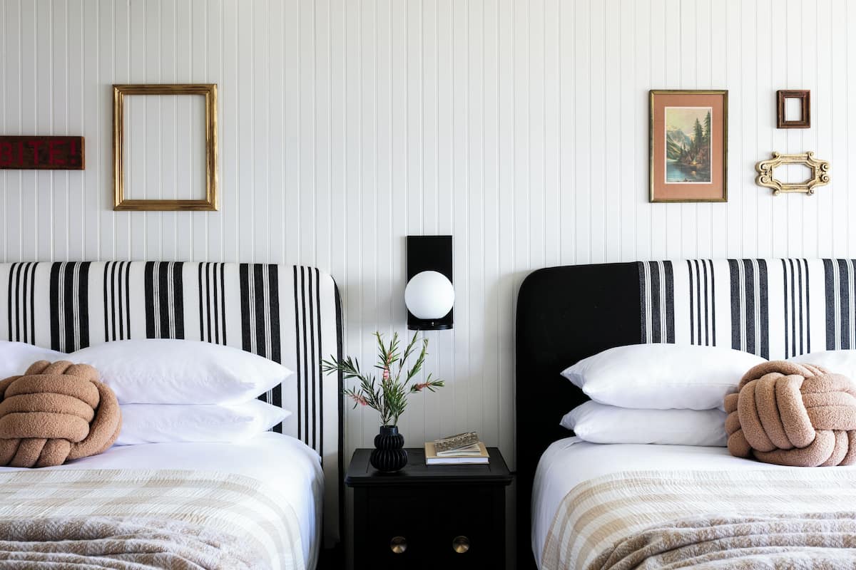 Ein Hotelzimmer mit zwei Betten, jeweils mit gestreifter Bettwäsche und einem einzelnen, braunen Zierkissen. Ein Nachttisch mit Lampe und einem kleinen Pflanzenarrangement steht zwischen den Betten.