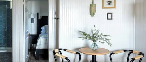 Une pièce comprenant un lit, une petite table avec un vase, deux chaises et une porte donnant sur une autre pièce.