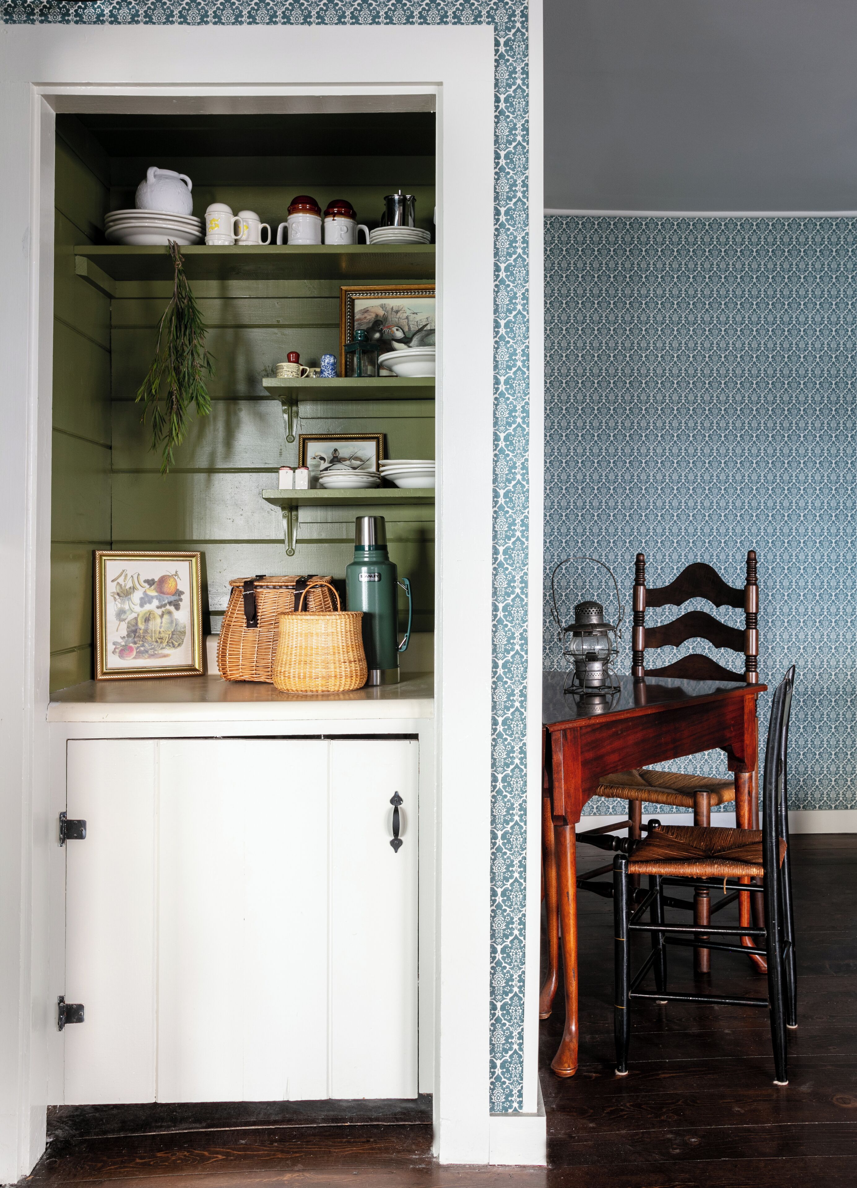 Una cocina con un armario blanco, repisas con vajilla, una cesta de mimbre y una silla de madera.