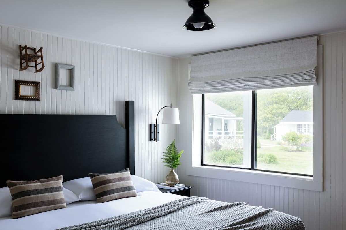Ein Schlafzimmer mit einem großen Bett, einer Nachttischlampe, einem Fenster mit Blick auf ein Haus und einer Deckenleuchte.