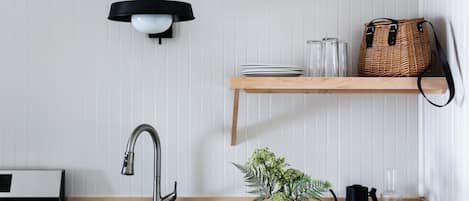 A kitchen with a stainless steel sink, a black wall-mounted light, a wooden shelf with glassware and a wicker basket, and a potted plant on the countertop.