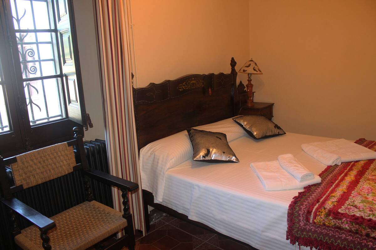 A bedroom with a wooden bed, a chair, a nightstand with a lamp, and a window with curtains.