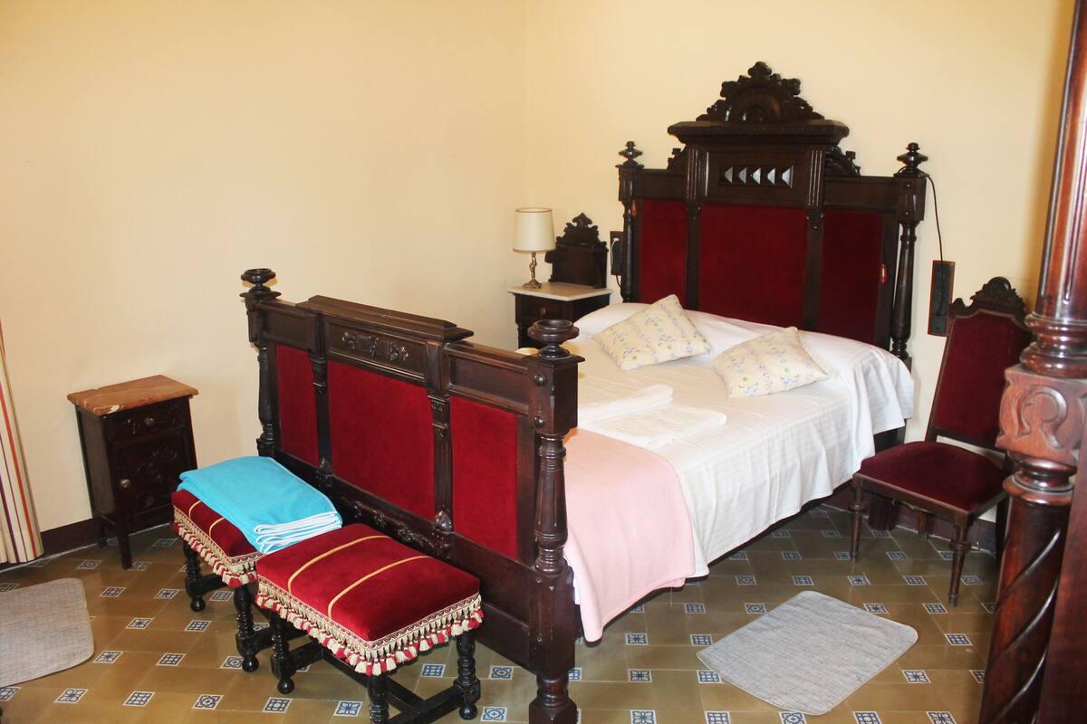 A traditional four-poster bed with a red headboard, a bedside table with a lamp, a wooden bench with a blue cover, and a red chair.