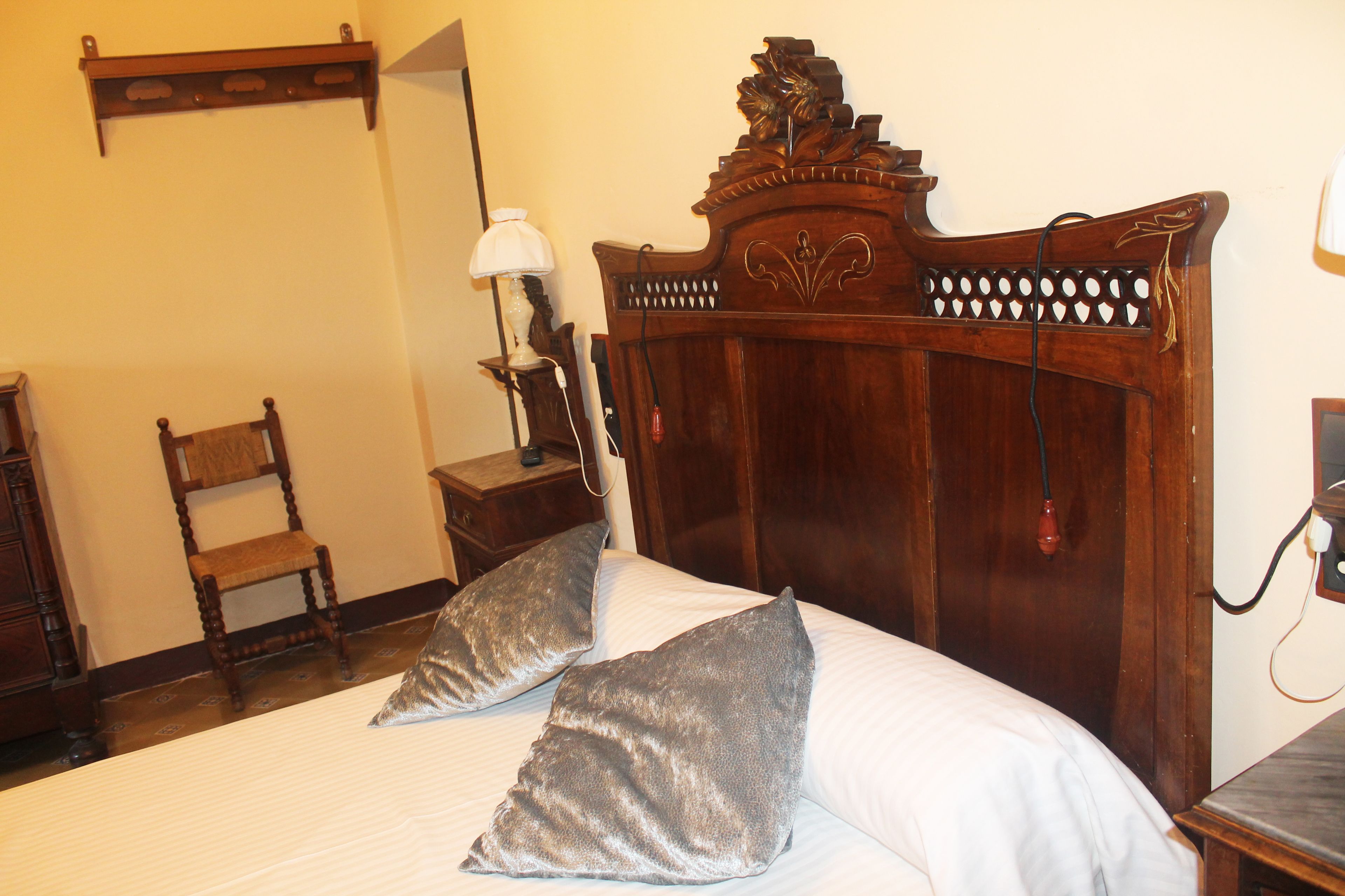 Una cama bien tendida con dos almohadas, un cabecero de madera, una mesita de noche con una lámpara y una silla en una habitación con paredes lisas.