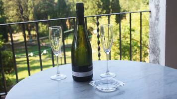 A bottle of wine and two glasses on a round table with a view of a garden and a balcony railing.
