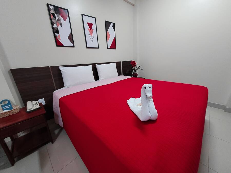 Una habitación de hotel con una colcha roja, una toalla blanca con forma de cisne, una mesita de noche de madera con un teléfono y una pared con tres cuadros.