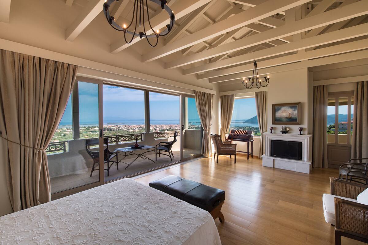 A spacious bedroom with a large bed, a balcony with a view, a fireplace, and a dining area.