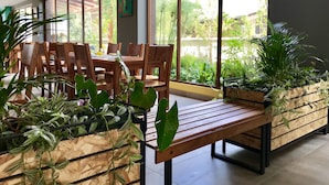 A modern dining area with wooden tables and chairs, large windows with a view of greenery, and potted plants. - Ilalo Garden Hotel & Restaurant (Tumbaco)