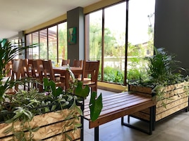 A modern dining area with wooden tables and chairs, large windows with a view of greenery, and potted plants.