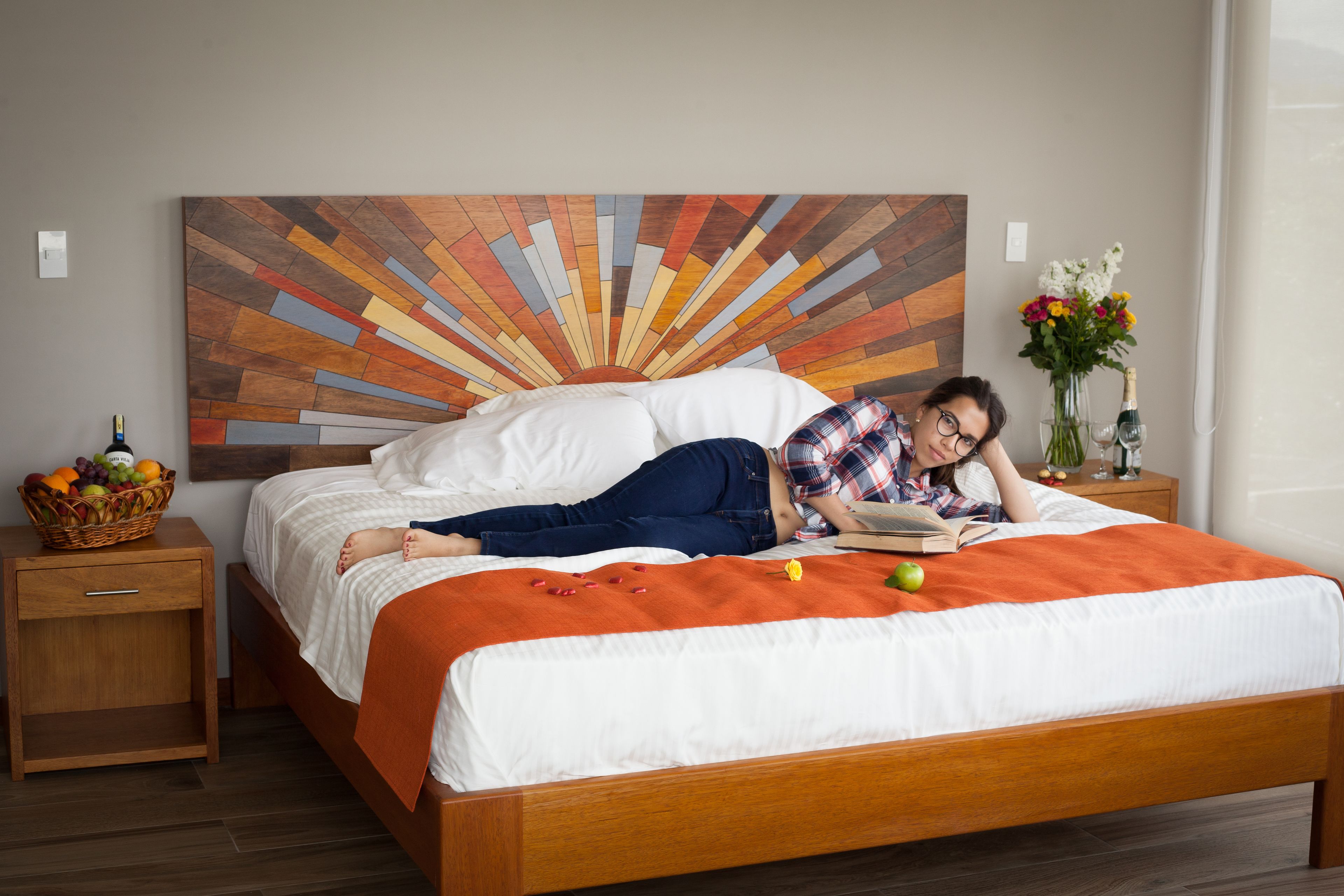 Una cama con cabecera de madera, una colcha blanca y una manta naranja. Hay una cesta de fruta sobre una mesita de noche junto a la cama.