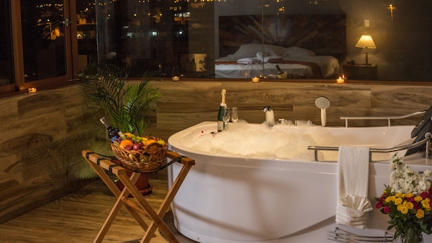 A modern bathroom with a large bathtub, a wooden floor, a wicker basket with fruits, and a city view through large windows. - Ilalo Garden Hotel & Restaurant (Tumbaco)
