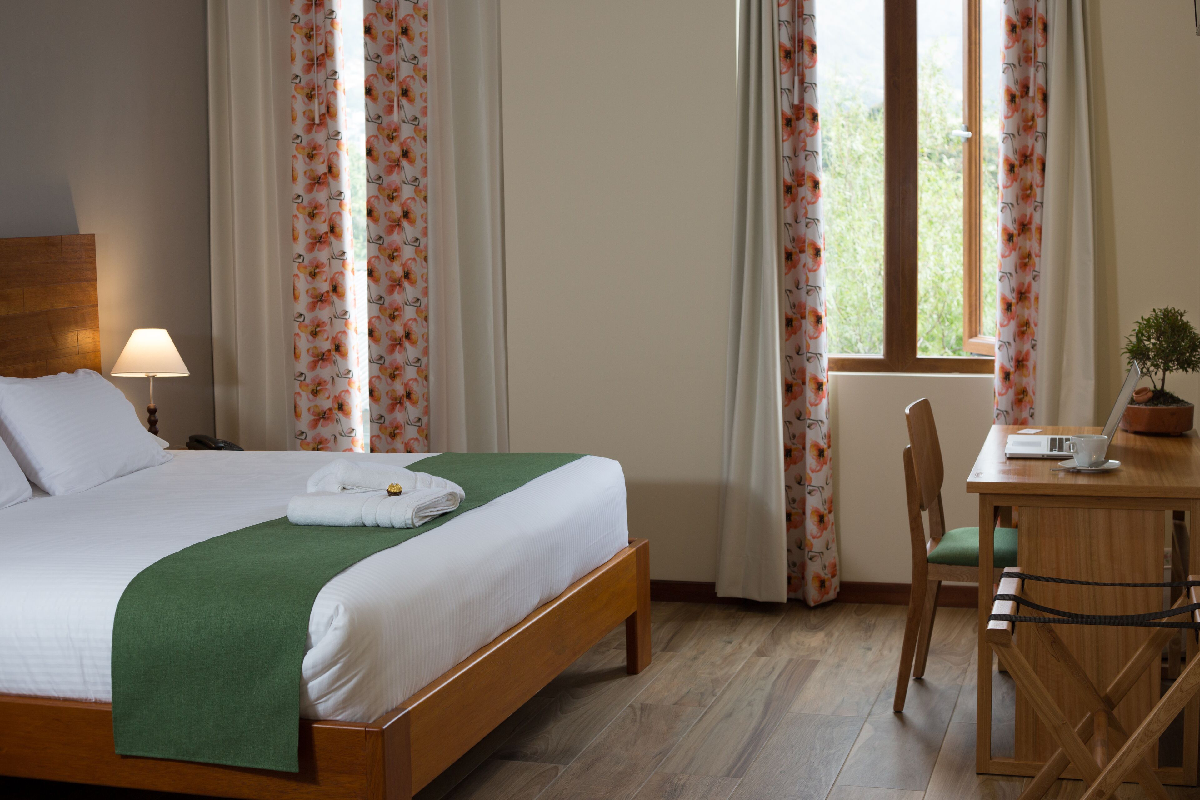 A neatly made bed with a green blanket, a wooden nightstand with a lamp, a desk with a laptop, and a chair.