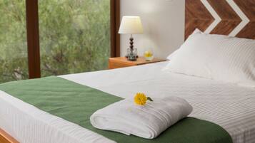 A neatly made bed with a green throw, a white towel, and a yellow flower. There is a lamp on a wooden nightstand beside the bed.