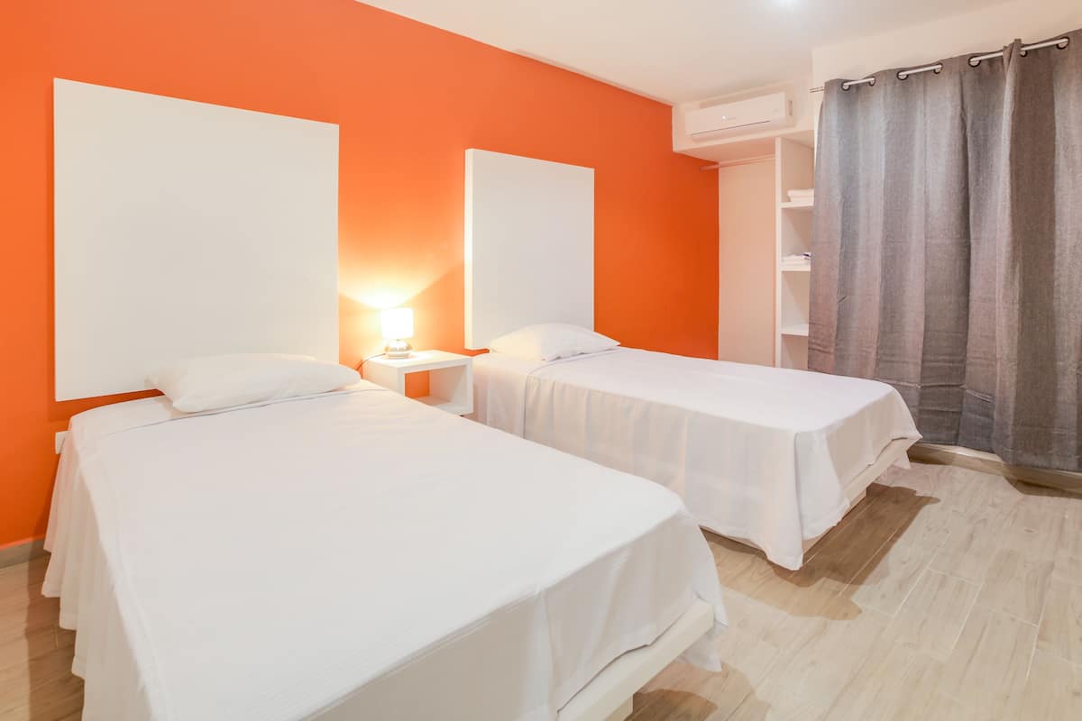 A hotel room with two single beds, a nightstand with a lamp, an air conditioning unit, and a grey curtain.
