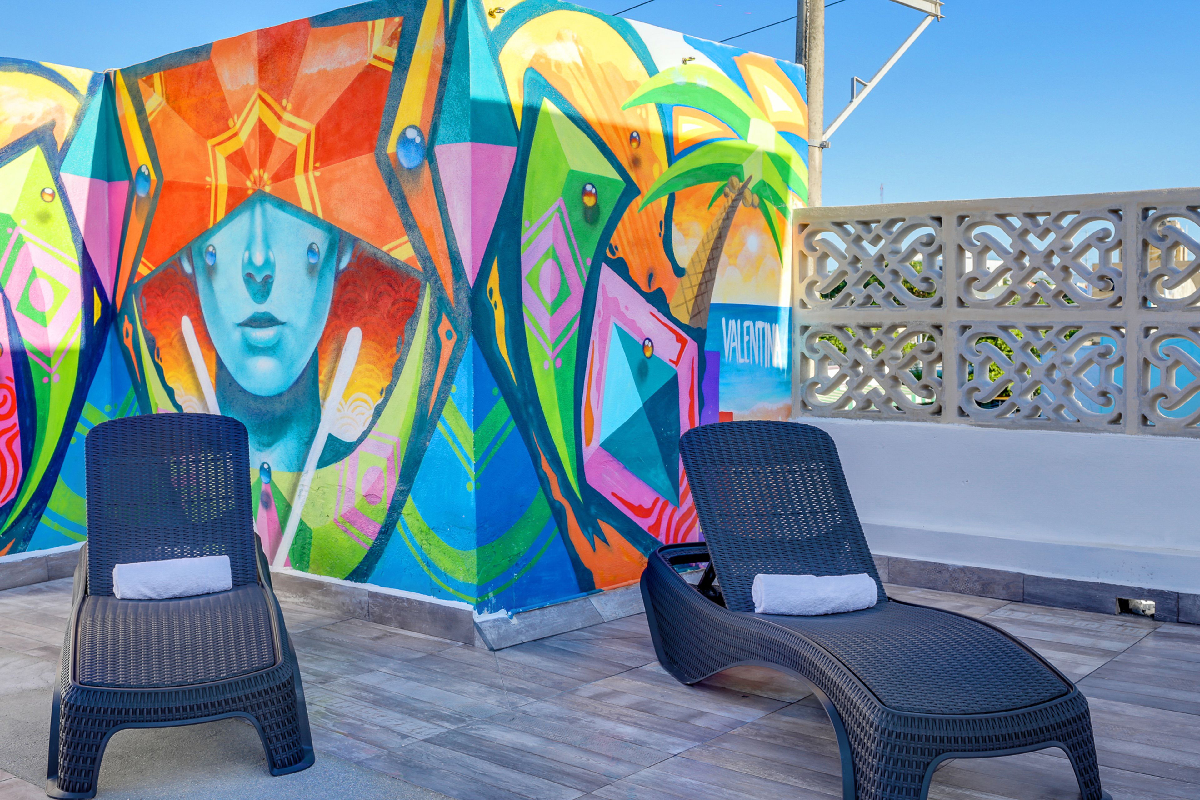 Deux fauteuils de jardin en osier, dotés de coussins blancs, sont placés sur une terrasse carrelée, avec une fresque colorée en arrière-plan.