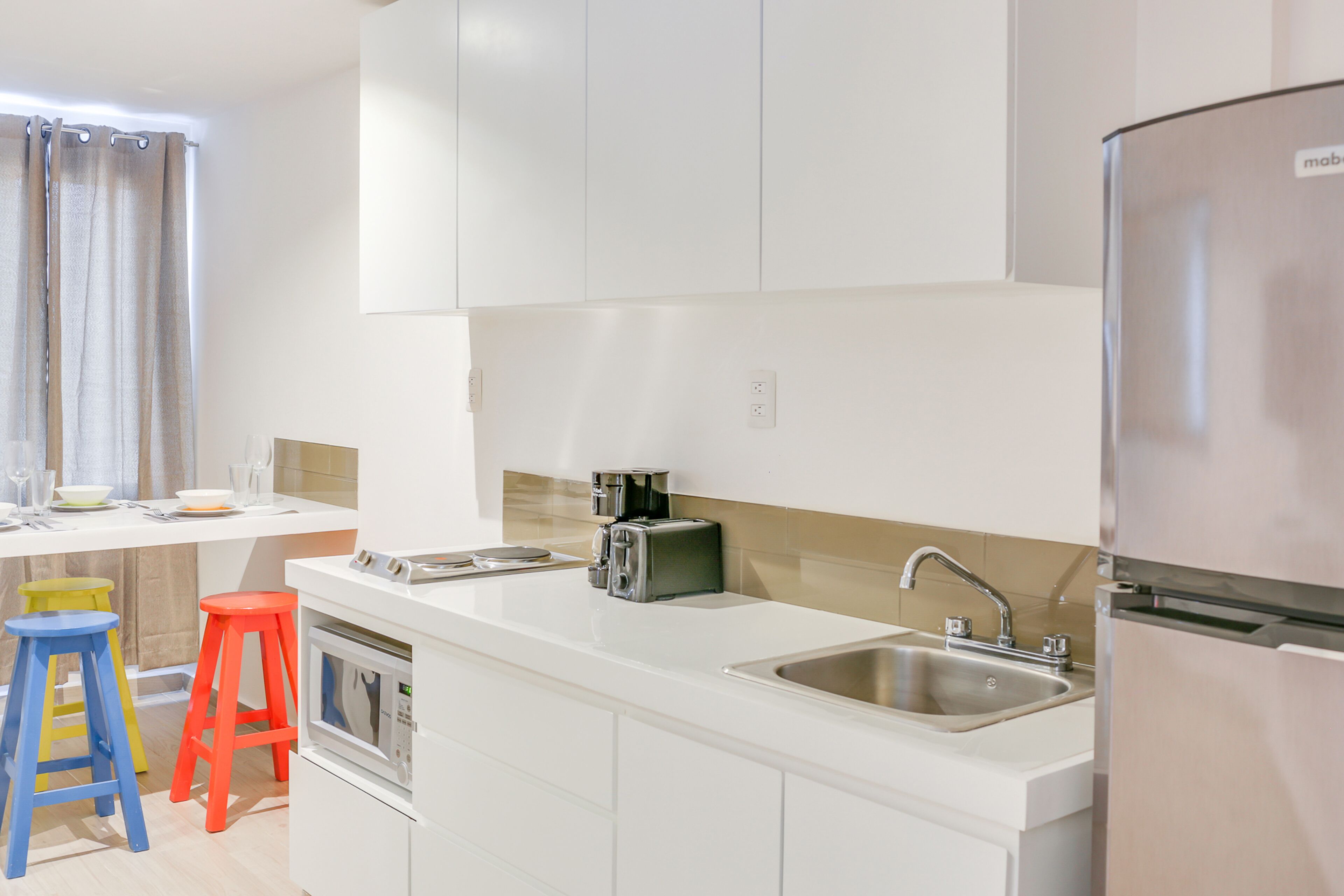 A modern kitchen with white cabinets, a stainless steel sink, a coffee maker, and two colorful stools.
