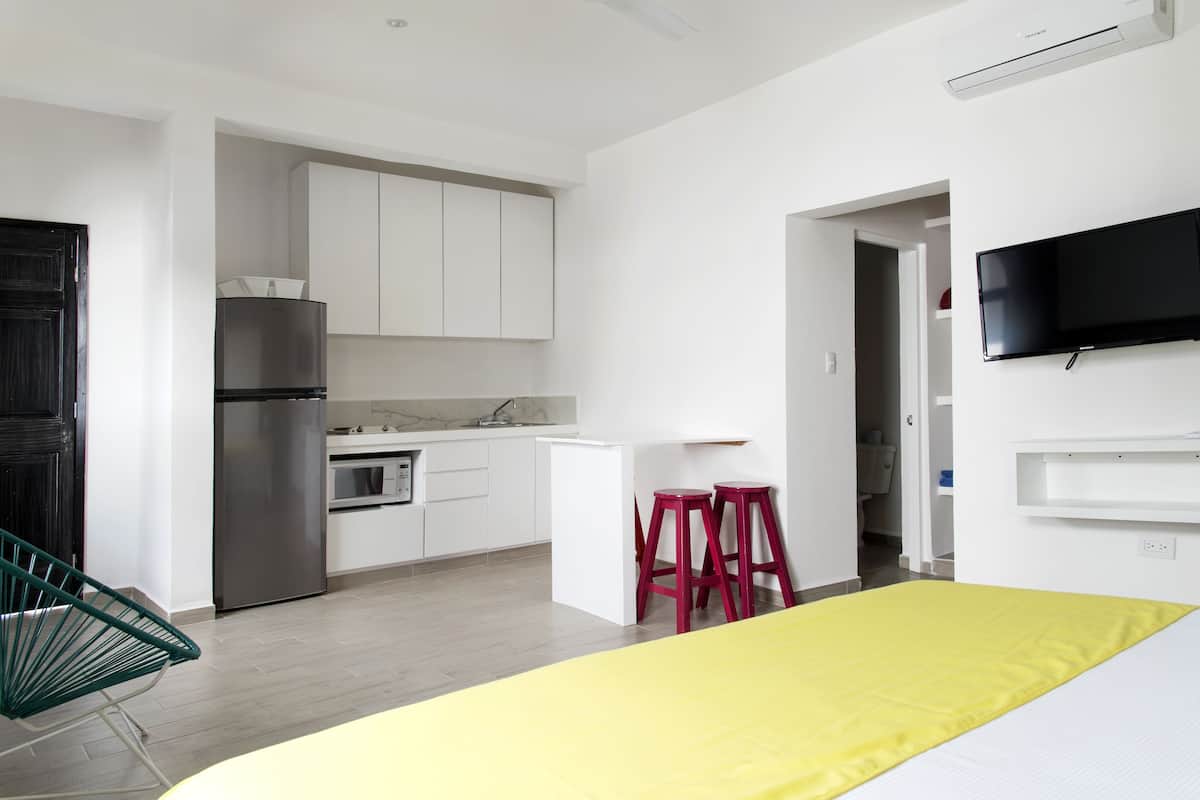 A modern kitchen with white cabinets, a stainless steel refrigerator, and a microwave. There is a yellow bed with a white mattress, a red stool, and a green chair.