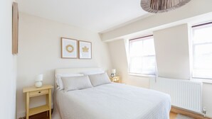A bedroom with a large bed, a bedside table with a lamp, a nightstand, a radiator, and two framed pictures on the wall. - Lovely Apartment Angel (London)
