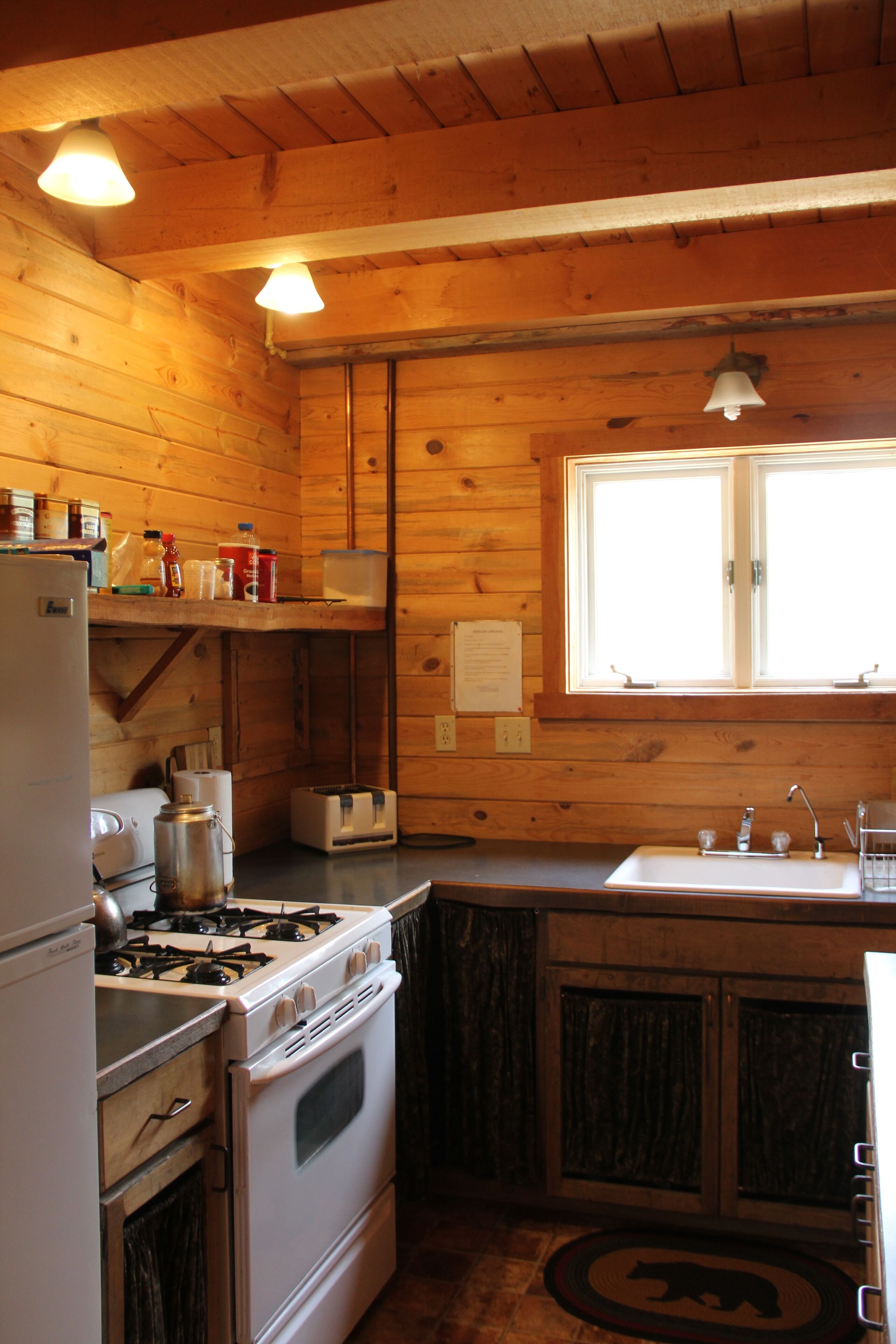 A compact kitchen with wooden walls, a gas stove, a refrigerator, a sink, and a window with a view.