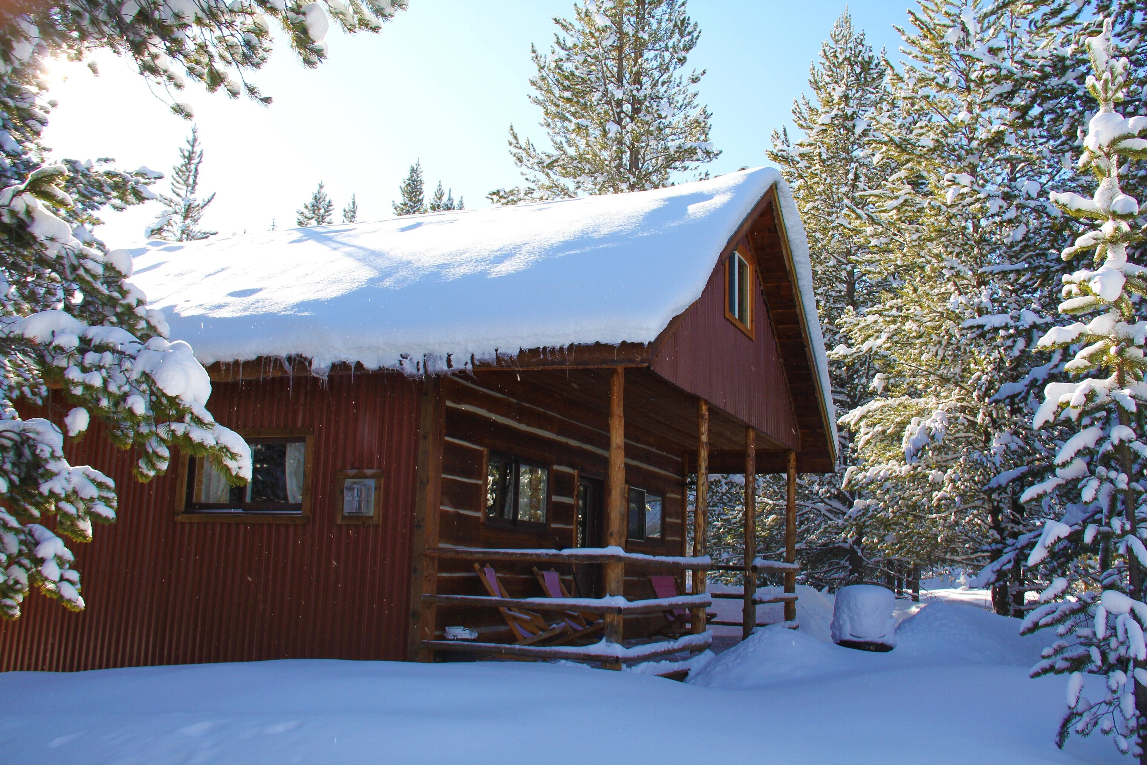 A wooden cabin with a snow-covered roof and porch, surrounded by snow-laden trees.