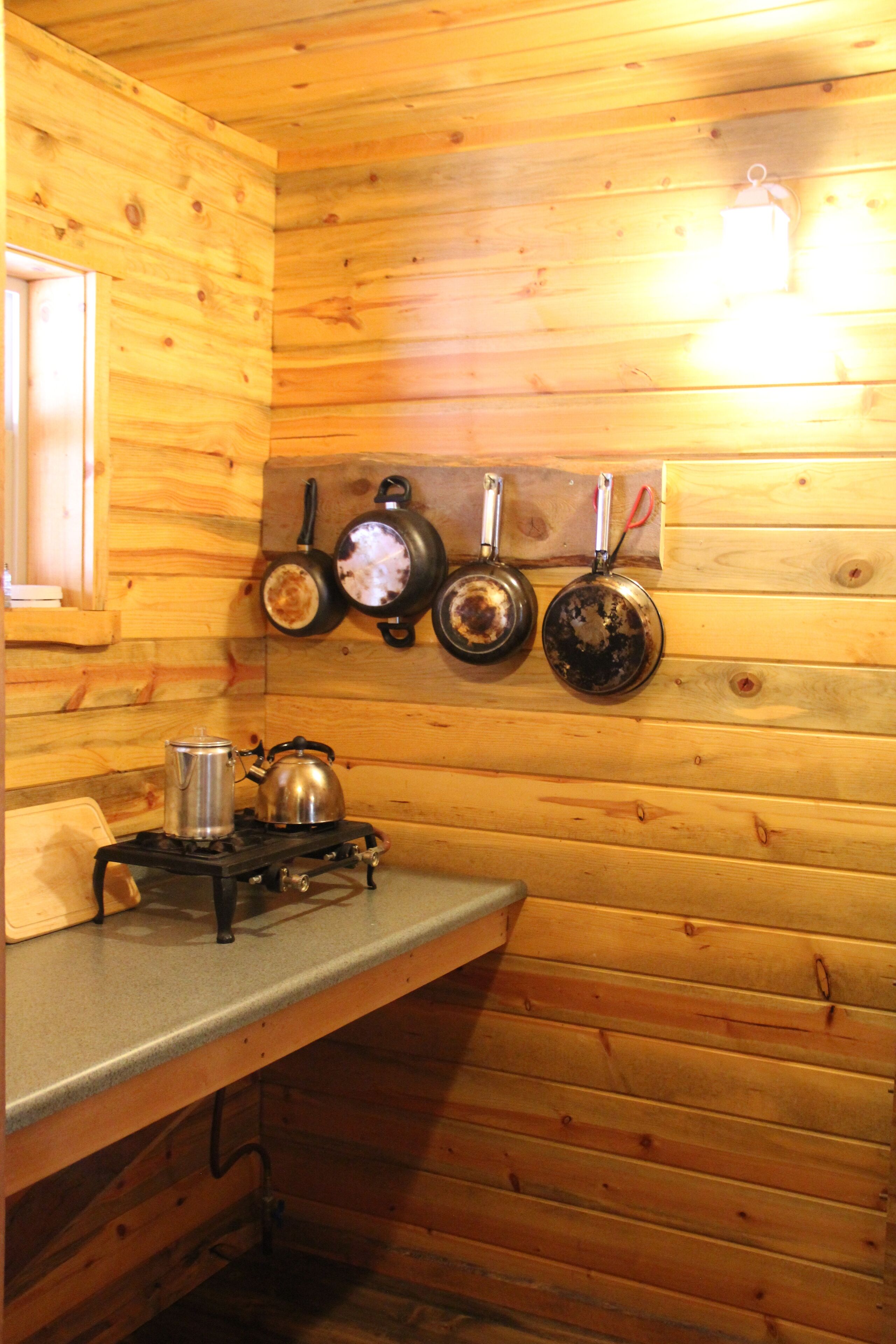A wooden cabin interior with a kitchen area featuring a stove, kettle, and pots hanging on the wall.