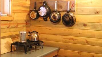 A wooden cabin interior with a kitchen area featuring a stove, kettle, and pots hanging on the wall.