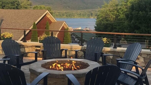 En bålplads omgivet af Adirondack-stole på en terrasse med udsigt over en sø og skovklædte bakker.