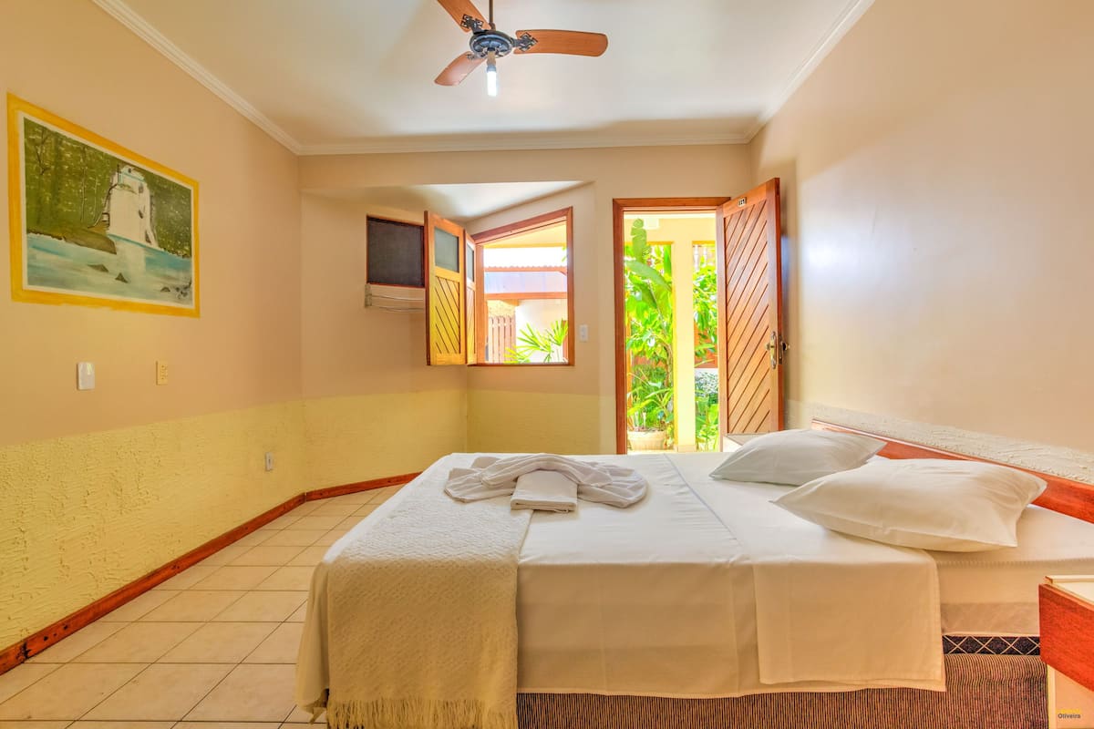 A hotel room with a bed, a ceiling fan, a painting, and a view of the outdoors.