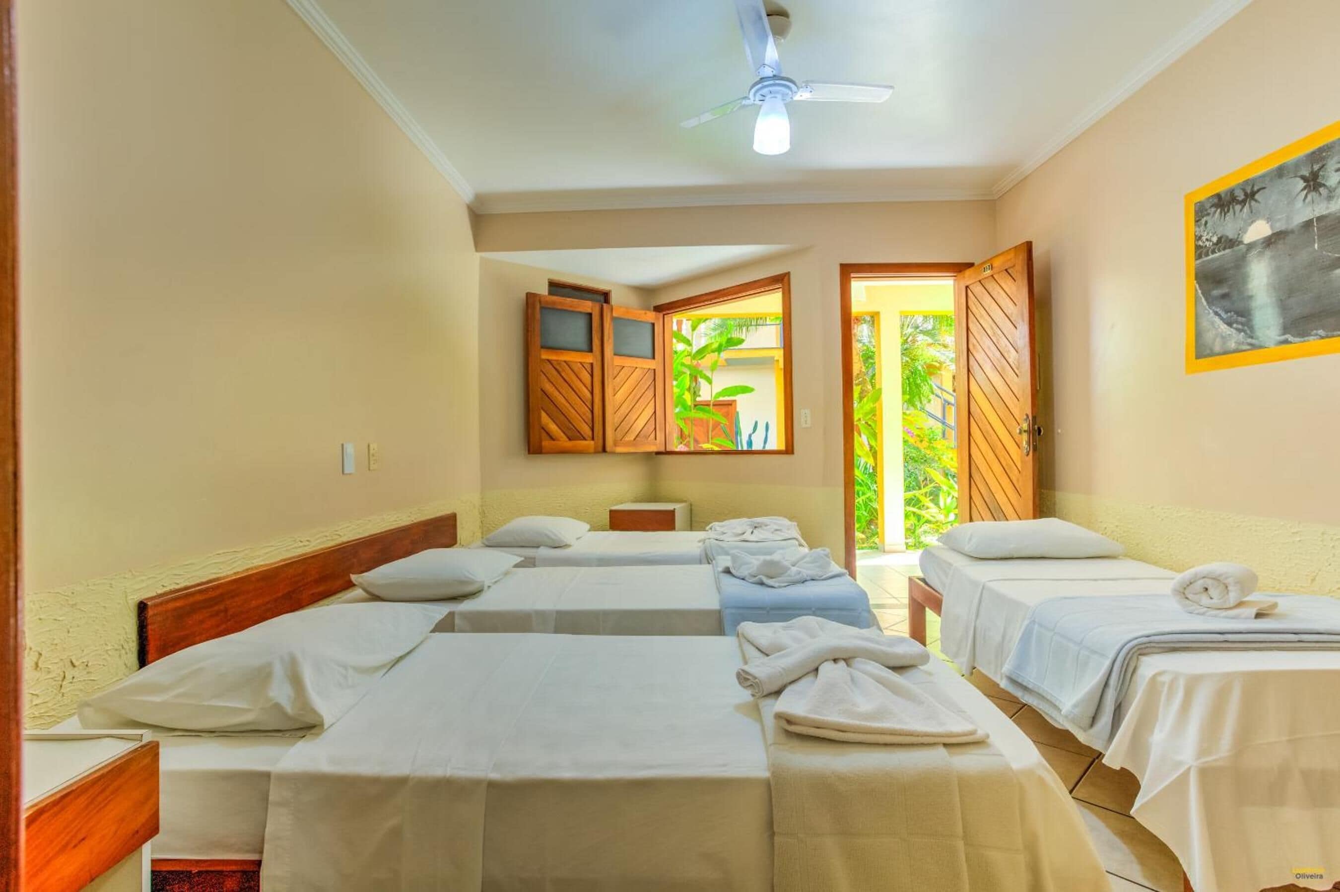 Habitación de hotel con cuatro camas individuales, ventilador de techo, un cuadro y vistas al exterior con vegetación.