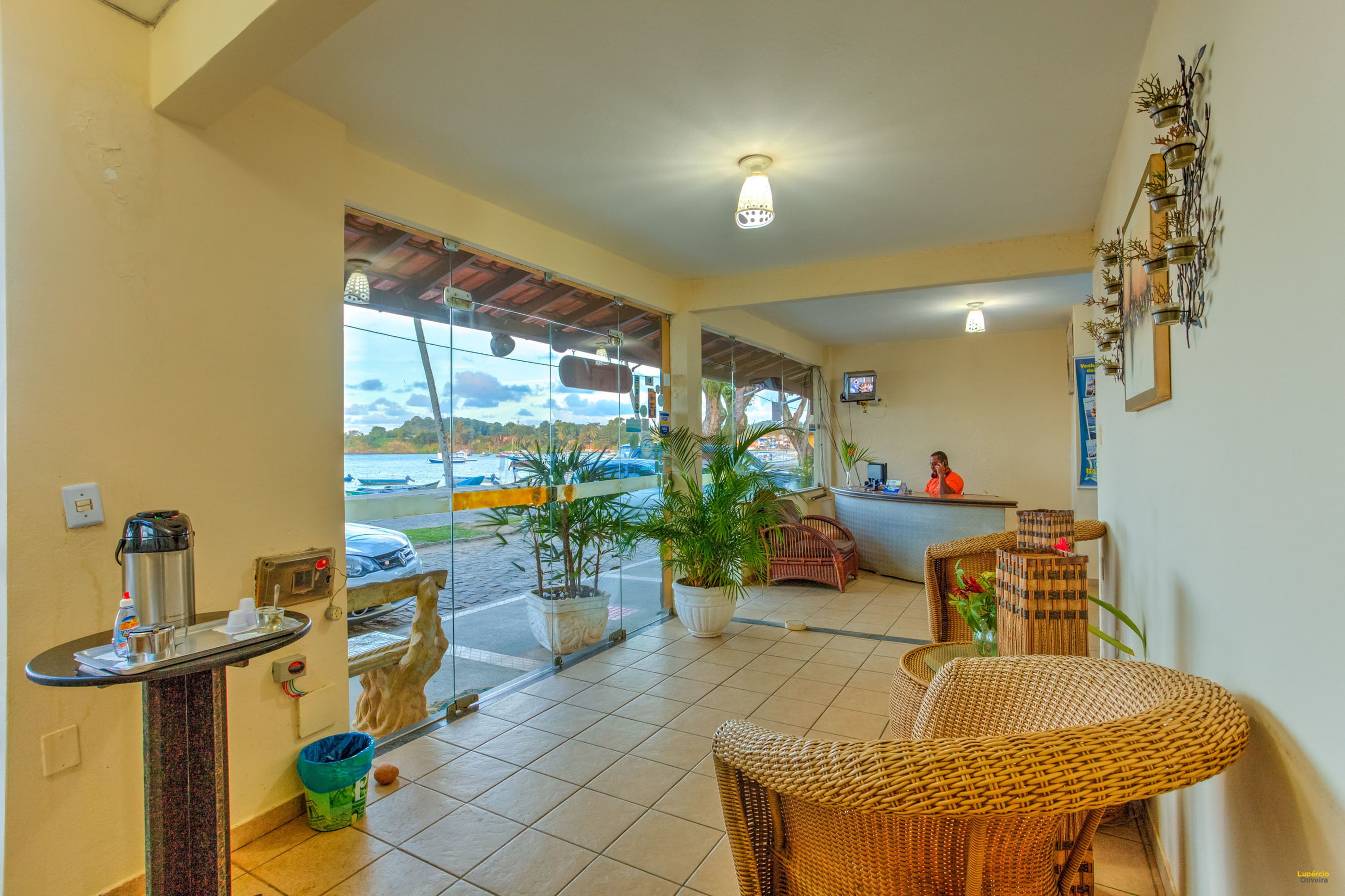 A hotel lobby with a reception desk, wicker furniture, potted plants, and a view of the street outside.