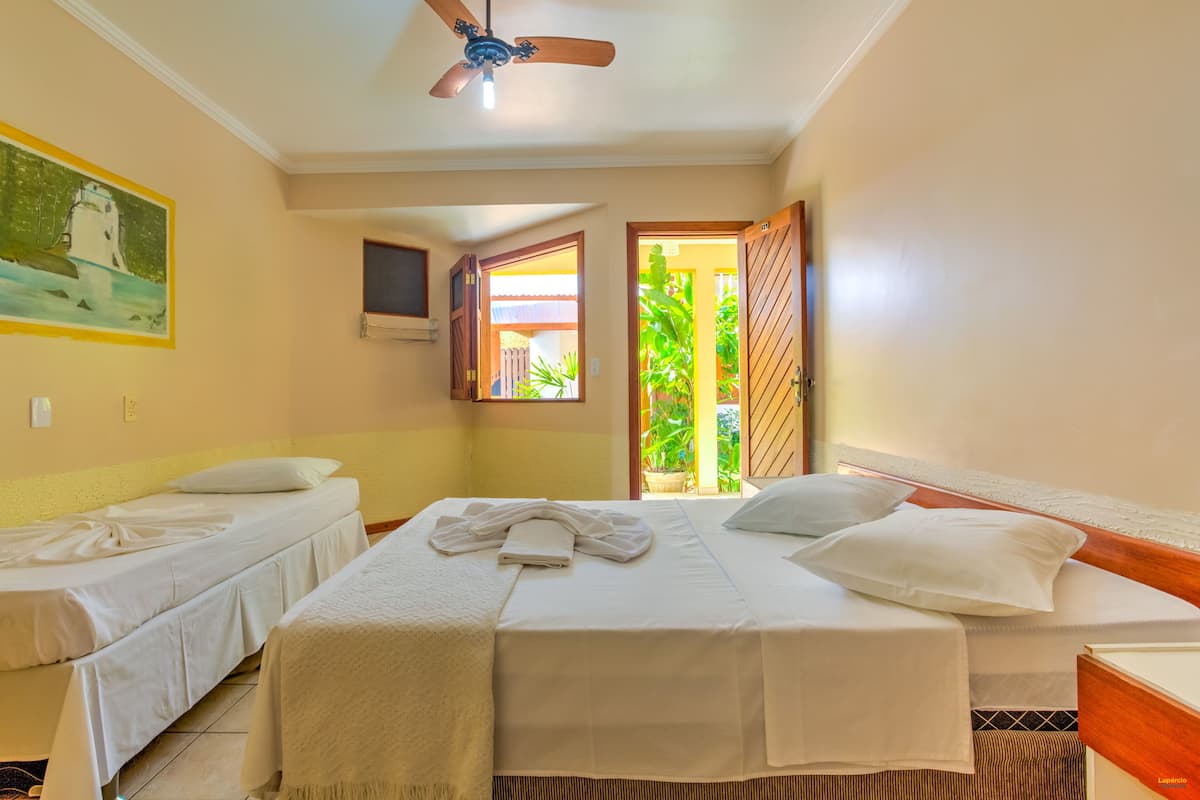 A hotel room with two single beds, a ceiling fan, a painting on the wall, and a view of greenery through an open door.