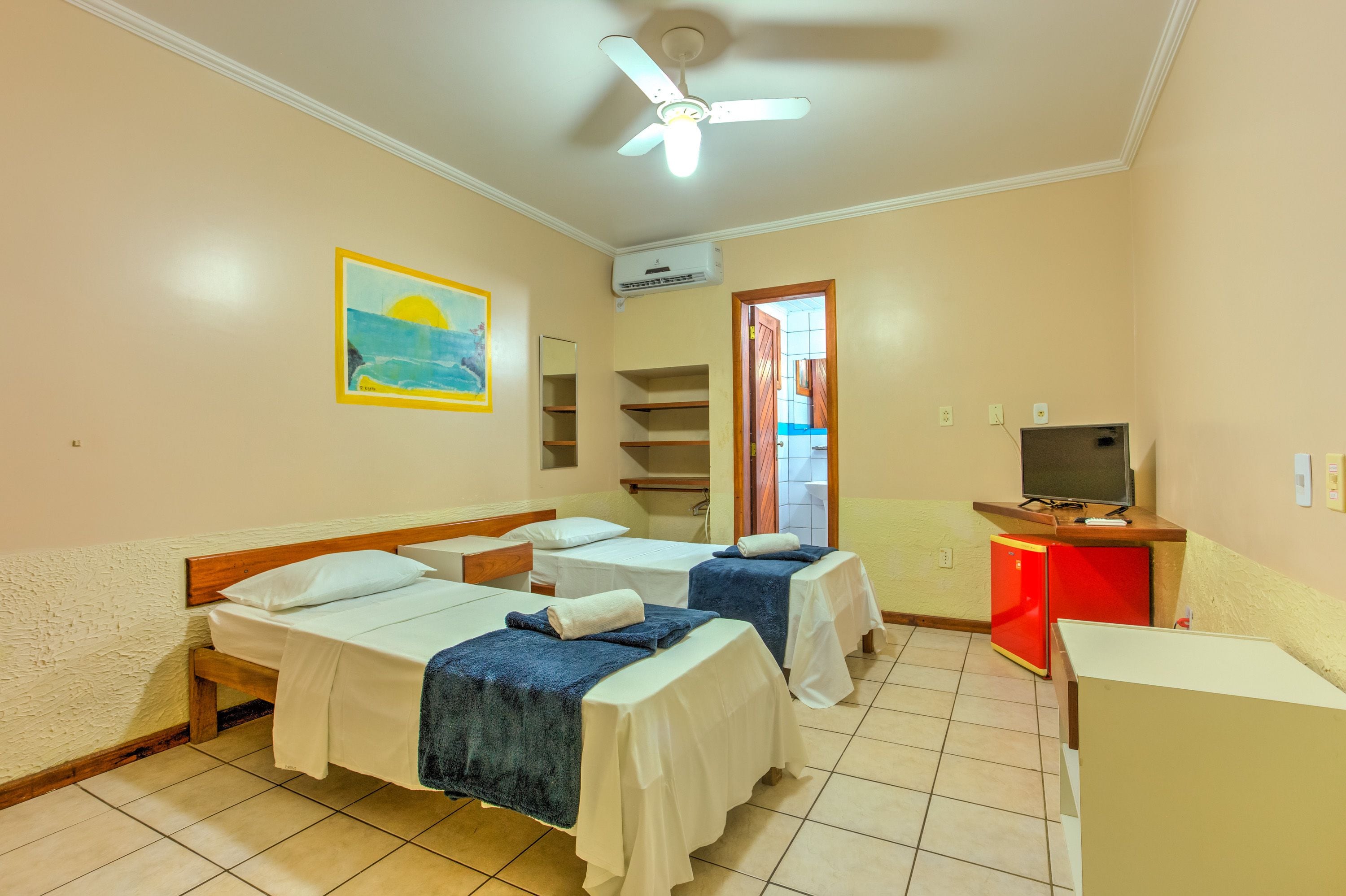 Habitación de hotel con dos camas, ventilador de techo, aire acondicionado, televisor y un escritorio pequeño.
