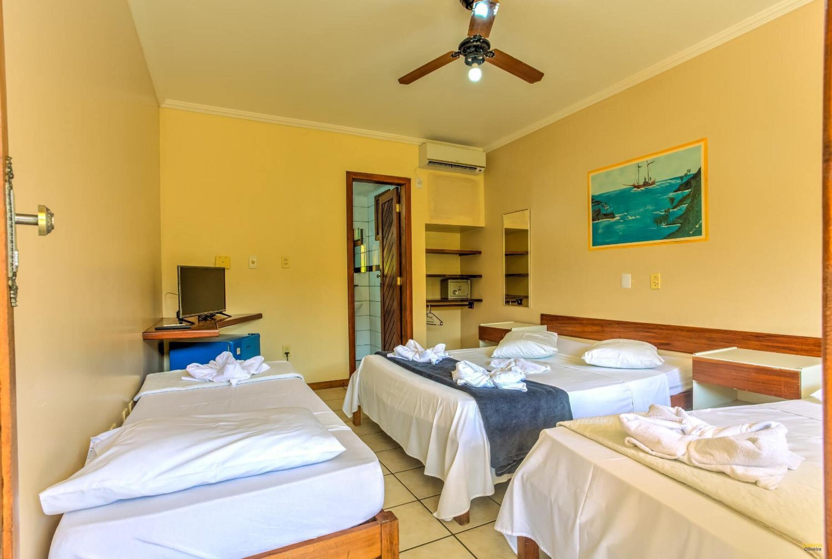 A hotel room with three beds, a ceiling fan, a painting of a coastal scene, and a television on a desk.