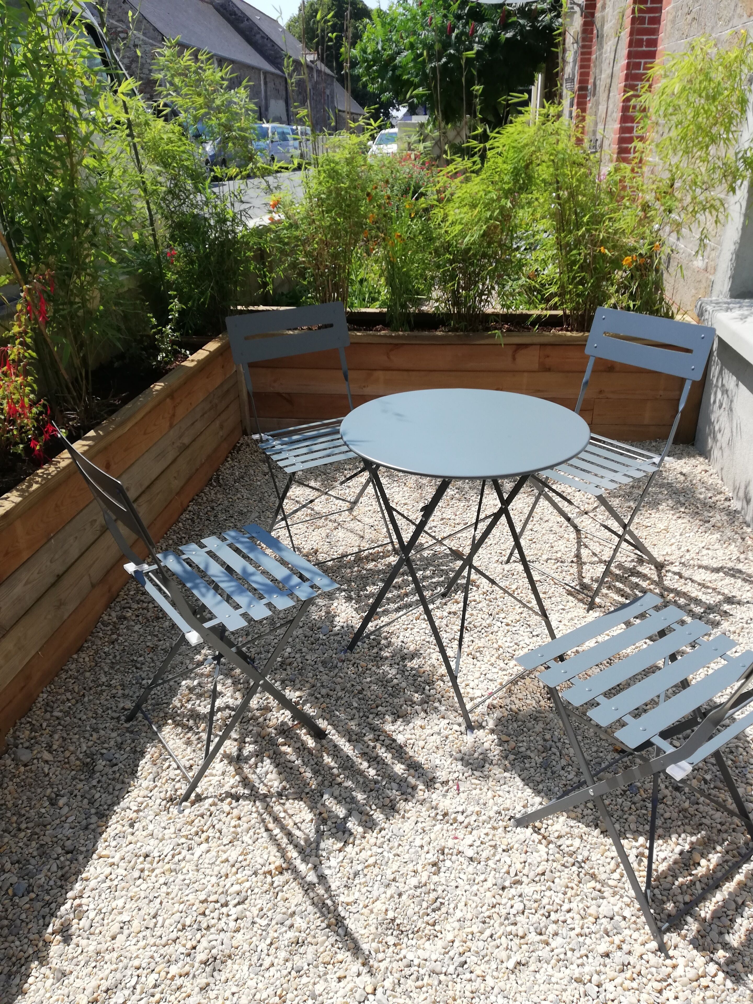 Une terrasse avec une table ronde et quatre chaises, entourée d’un jardinier en bois et d’un sol recouvert de gravier.