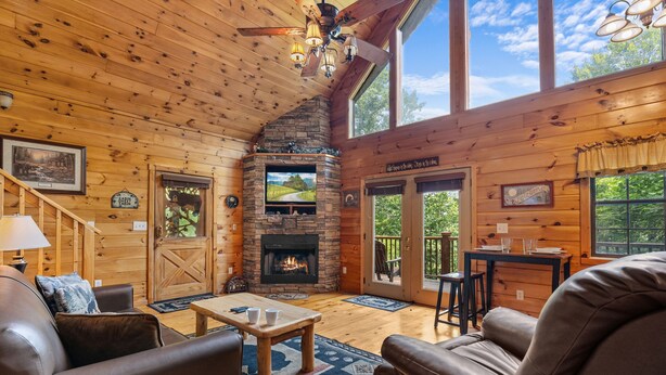A cozy cabin interior with a fireplace, wooden walls, leather furniture, and large windows offering a view of the outdoors.