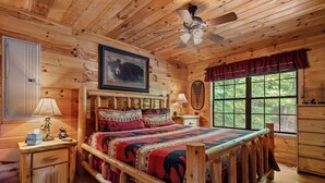 A wooden cabin bedroom with a bed, a ceiling fan, a window with a view of trees, and a wooden dresser.