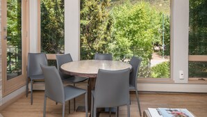 A modern dining area with a round table and six chairs, large windows offering a view of trees and hills, and a book titled 'The Stylish Life Skiing' on a small table. - Gateway Plaza R4 2 Bedroom Condo by RedAwning (Vail)
