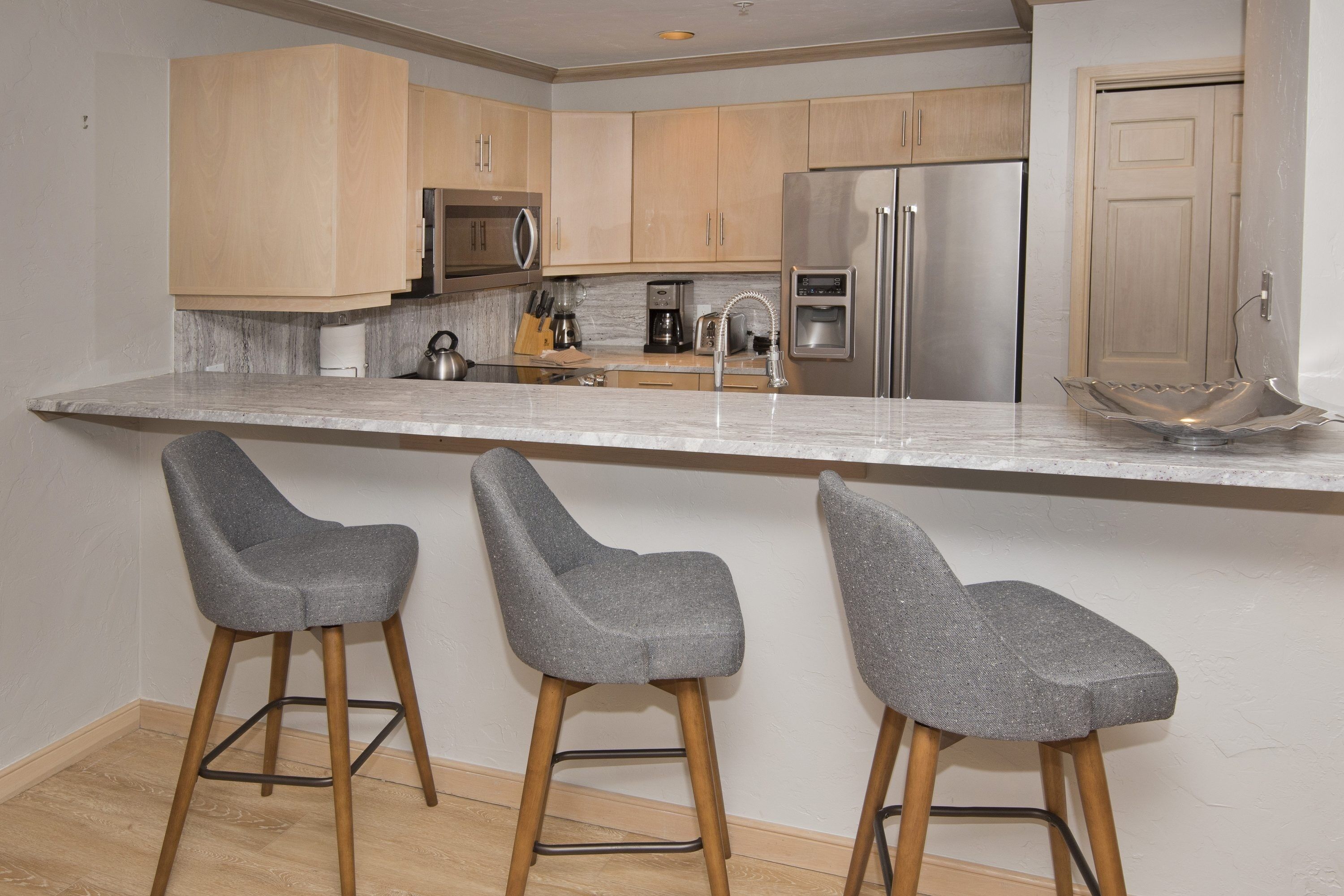 A modern kitchen with wooden cabinets, stainless steel appliances, and a marble countertop. There are three grey bar stools with wooden legs.