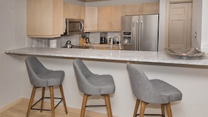 A modern kitchen with wooden cabinets, stainless steel appliances, and a marble countertop. There are three grey bar stools with wooden legs. - Gateway Plaza R4 2 Bedroom Condo by RedAwning (Vail)