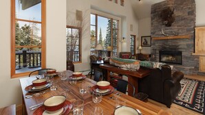 Un comedor con una gran mesa de madera dispuesta para una comida, una chimenea con pared de piedra y vistas a árboles desde el exterior.