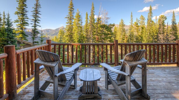 Una terraza de madera con dos sillas Adirondack y una pequeña mesa redonda, con vistas a un paisaje montañoso cubierto de bosque.
