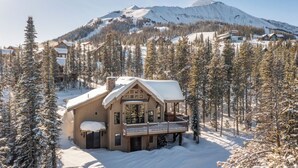 Una cabaña de madera con balcón, rodeada de árboles cubiertos de nieve y una montaña al fondo.