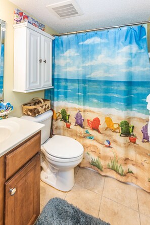 Ein Badezimmer mit Strandmotiven an der Duschvorhang, einer weißen Toilette, einem Holzschrank und einem blauen Teppich.