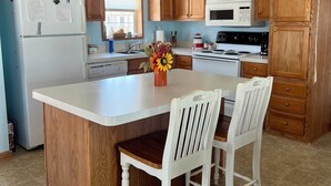 A kitchen with wooden cabinets, a white refrigerator, a microwave, and a stove. There is a wooden island with two white chairs and a vase with flowers. - Lake Front Cottage Perfect for Fisherman, Birders,  and Family Vacations. (Oak Harbor)