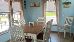 A dining room with a wooden table and white chairs, light blue walls, and a view of the outdoors through large windows. - Lake Front Cottage Perfect for Fisherman, Birders,  and Family Vacations. (Oak Harbor)