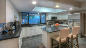A modern kitchen with white cabinets, a large island with bar stools, a built-in oven, and a large window overlooking a garden. - Family Home with Pool, Hot Tub & BBQ near Kierland (Scottsdale)