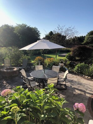 An outdoor patio with a round table and chairs, surrounded by lush greenery and a white umbrella.