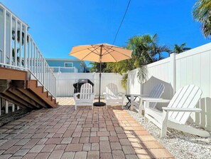 A patio with a brick floor, white Adirondack chairs, a patio umbrella, and a white fence.